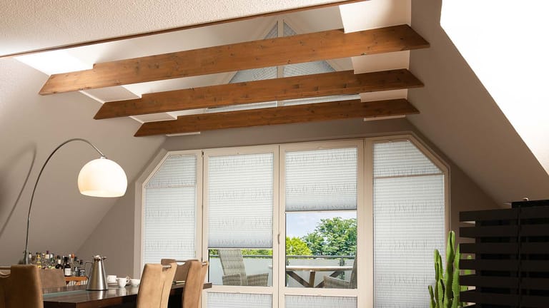 Ein Esszimmer mit einem Holztisch, beigen Stühlen, einer Stehlampe und großen Fenstern mit Jalousien. Freigelegte Balken und eine Topfpflanze sorgen für Dekoration.
